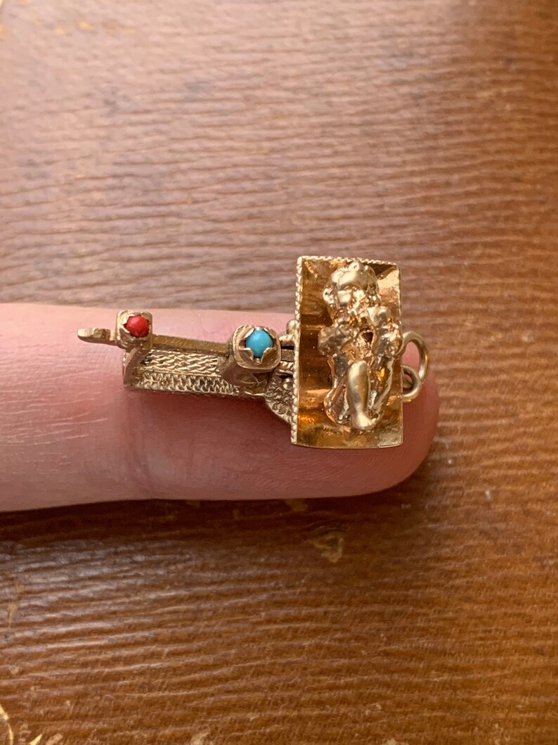 Op de afbeelding: Een goudkleurige bedel met een rechthoekig frame met een gebeeldhouwde figuur. De bedel heeft een kleine rode en turkooizen steen. De bedel zit op een vinger.