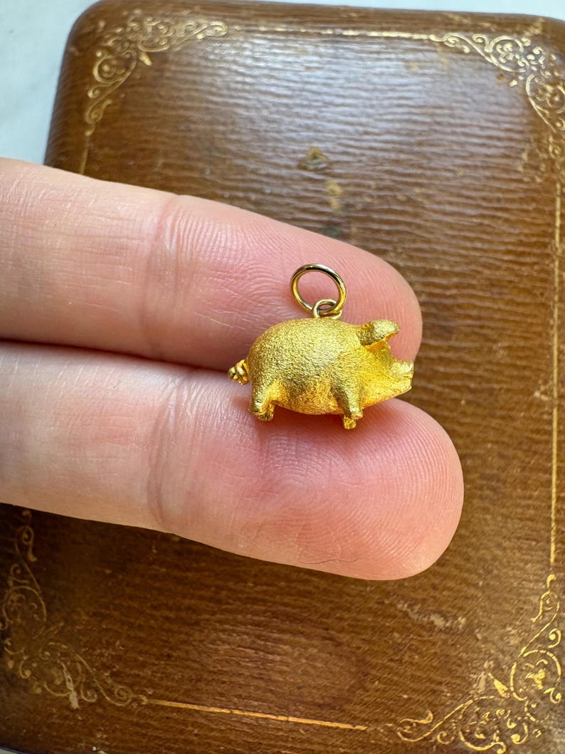 May include: A small, gold-colored pig charm with a textured surface. The charm has a loop at the top for attaching to a necklace or bracelet. The pig has small legs, a curly tail, and a detailed snout.