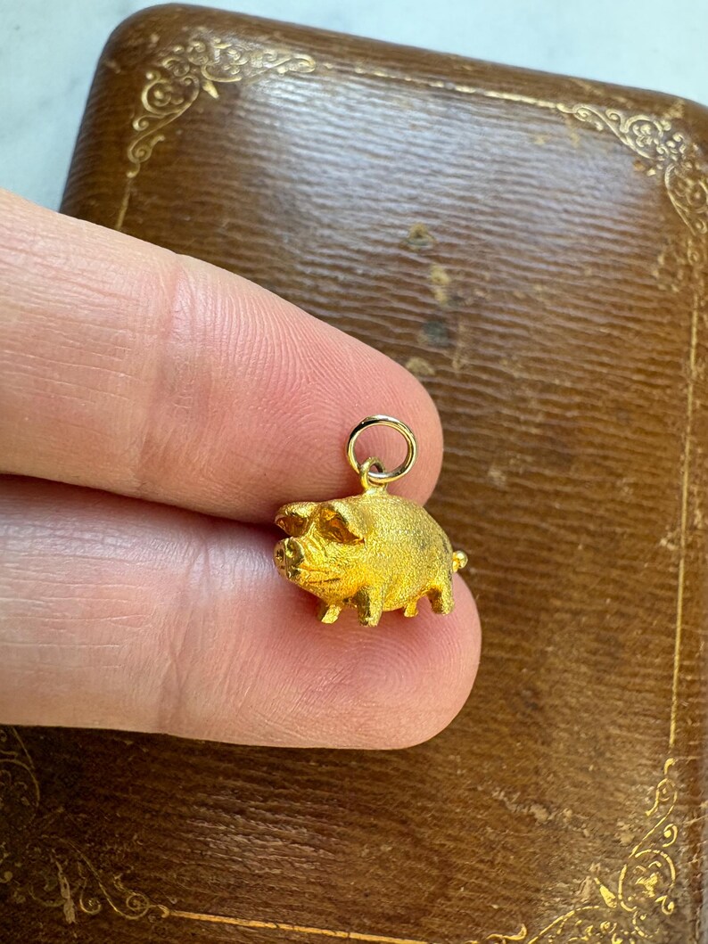 May include: A small, gold-colored pig charm with a textured surface. The charm is suspended from a small ring. The pig has visible legs, ears, and snout. The charm is held in a person's fingers.