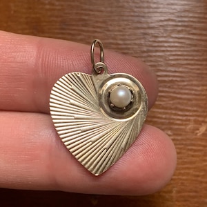 May include: A gold heart-shaped pendant with a pearl in the centre. The heart has a textured design with radiating lines.