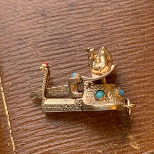 Op de afbeelding: Een goudkleurige metalen armbandsluiting met een gedetailleerd ontwerp. De sluiting heeft turquoisekleurige stenen en een rode steenaccent. De sluiting ligt op een bruin oppervlak.