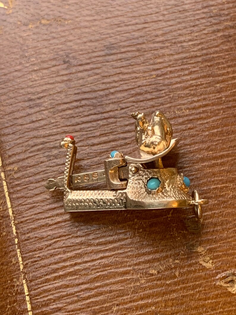 Op de afbeelding: Een goudkleurige metalen armbandsluiting met een gedetailleerd ontwerp. De sluiting heeft turquoisekleurige stenen en een rode steenaccent. De sluiting ligt op een bruin oppervlak.