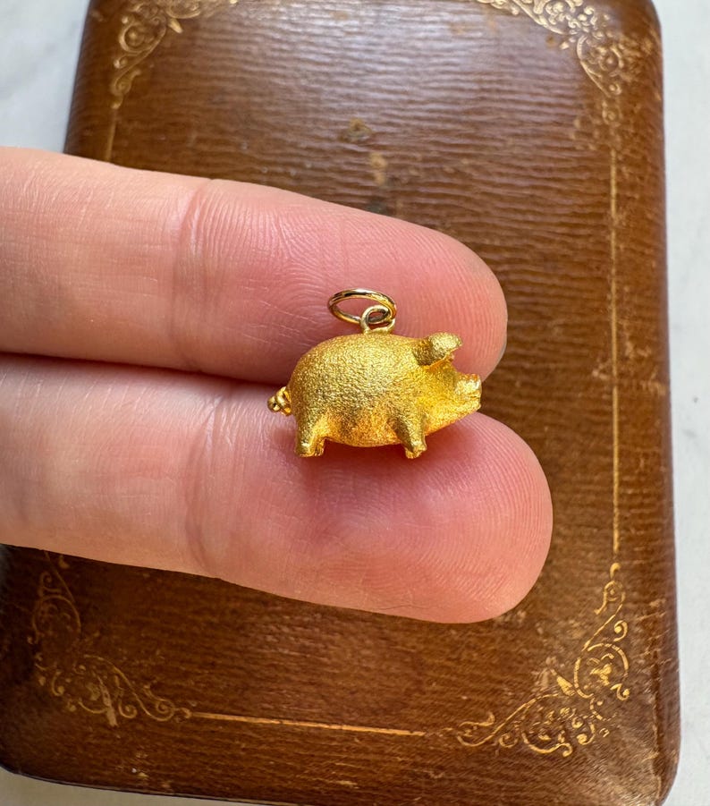 May include: A small, gold-colored pig charm with a textured surface. The charm is suspended from a small loop, suggesting it could be used as a pendant or charm. The pig is held between fingers against a brown background.