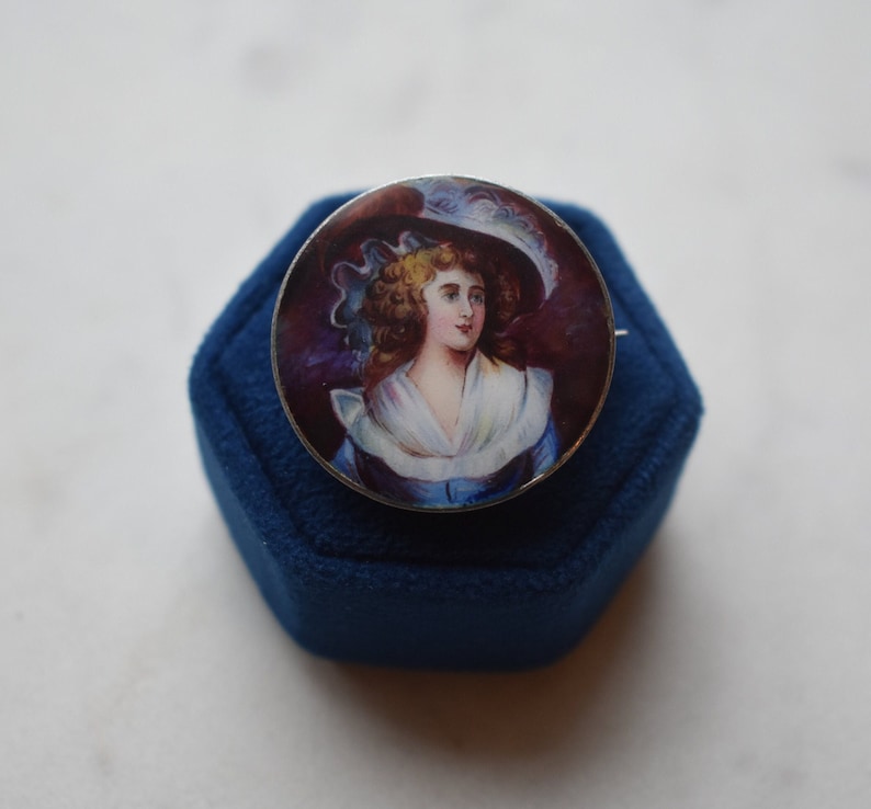 K&ouml;nnte beinhalten: Eine silberfarbene Brosche mit einem runden, gemalten Portr&auml;t einer Frau in einem blauen Kleid und einem wei&szlig;en Hut. Das Portr&auml;t ist vor einem dunkelvioletten Hintergrund platziert.