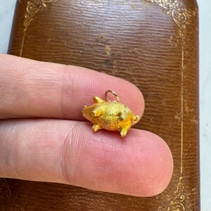 May include: A small, gold-colored pig charm with a textured surface. The charm has tiny legs, ears, and a curled tail. It is held against a brown, decorative box. The charm is approximately 0.5 inches in length.