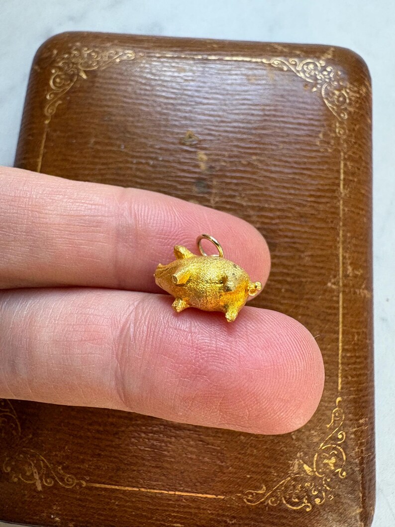 May include: A small, gold-colored pig charm with a textured surface. The charm has tiny legs, ears, and a curled tail. It is held against a brown, decorative box. The charm is approximately 0.5 inches in length.