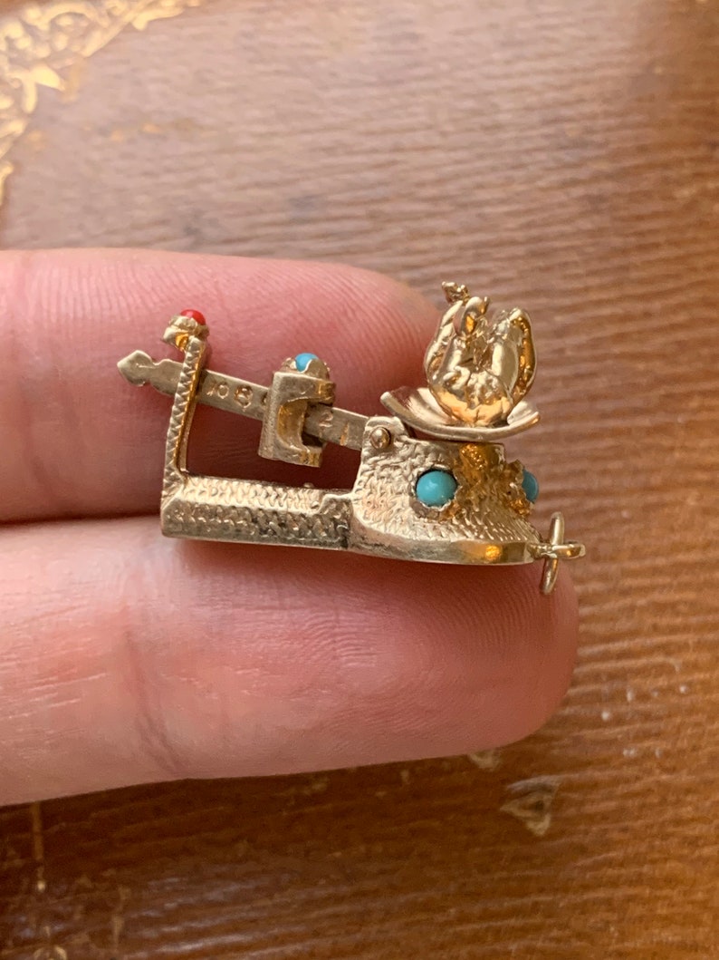 Op de afbeelding: Een goudkleurige bedel in de vorm van een weegschaal, met een kleine theepot erop. De bedel heeft turquoise en rode accenten. De weegschaal is gedetailleerd met ge&euml;tste lijnen en een klein kruisvormig handvat.