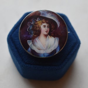K&ouml;nnte beinhalten: Eine silberne Brosche mit einem gemalten Portr&auml;t einer Frau in einem wei&szlig;en Hut und einem blauen Kleid. Das Portr&auml;t ist vor einem dunkelvioletten Hintergrund platziert.