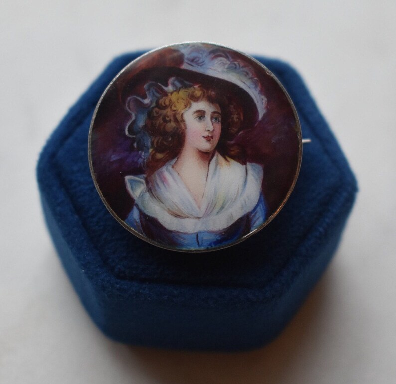 K&ouml;nnte beinhalten: Eine silberne Brosche mit einem gemalten Portr&auml;t einer Frau in einem wei&szlig;en Hut und einem blauen Kleid. Das Portr&auml;t ist vor einem dunkelvioletten Hintergrund platziert.