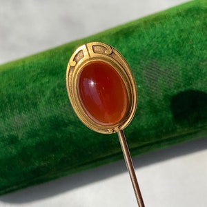 Könnte beinhalten: Eine goldfarbene Stecknadel mit einem ovalen Karneol-Stein, der in einen dekorativen Rahmen gefasst ist.