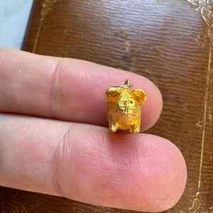 May include: A small, gold-colored pig charm with a loop for a necklace or bracelet. The pig has detailed features, including ears, snout, and legs. The charm is held between two fingers, with a brown, ornate box in the background.
