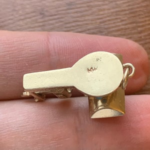 Op de afbeelding: Een kleine, goudkleurige fluitjesbedel met een afgeronde bovenkant en een cilindrische basis. De bedel heeft een lus om aan een ketting of armband te bevestigen. De fluit wordt in de hand van een persoon gehouden.