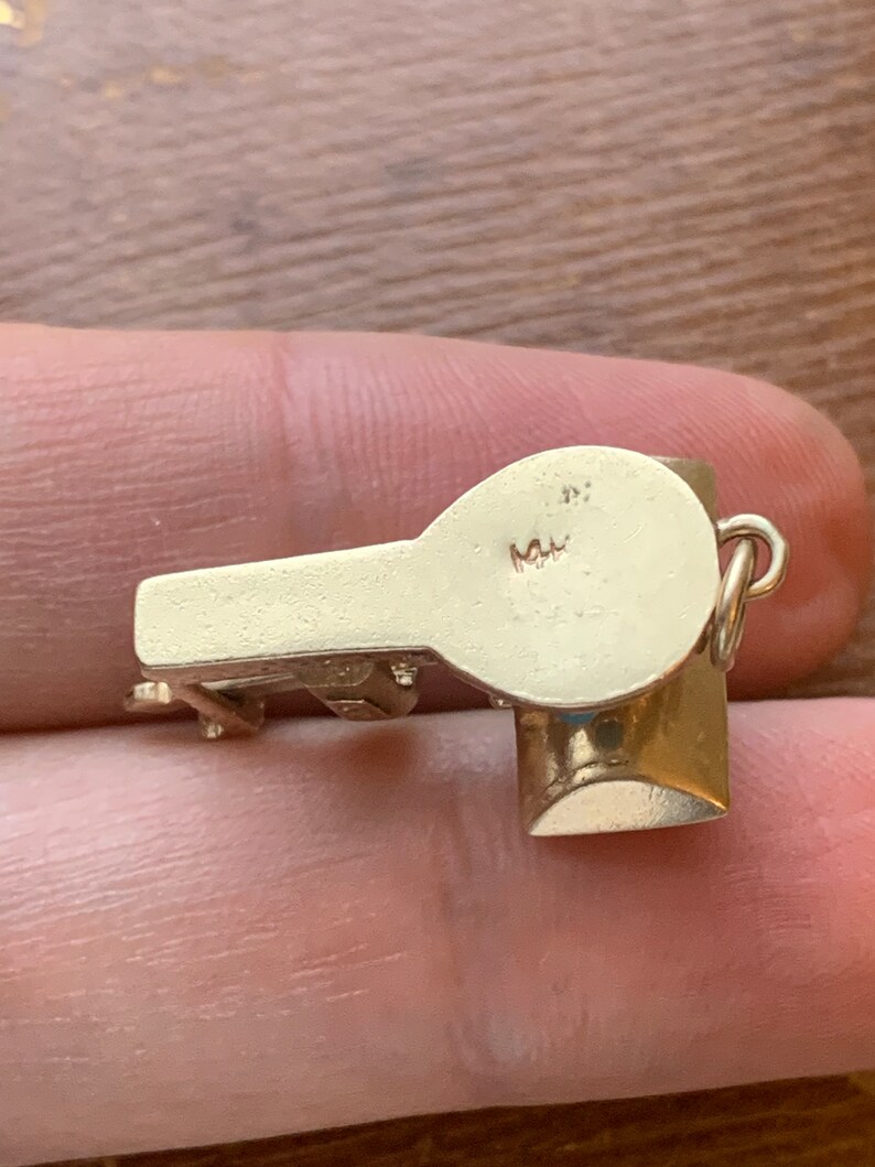 Op de afbeelding: Een kleine, goudkleurige fluitjesbedel met een afgeronde bovenkant en een cilindrische basis. De bedel heeft een lus om aan een ketting of armband te bevestigen. De fluit wordt in de hand van een persoon gehouden.