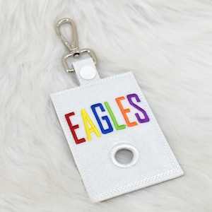 May include: White, rectangular pet waste bag holder with the word "EAGLES" embroidered in rainbow colors. It has a silver-colored metal clip and a circular opening for dispensing bags.