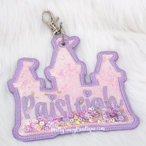 May include: Purple and pink castle-shaped keychain with the name "Paisley" spelled out in glitter. The keychain is decorated with colorful sequins.