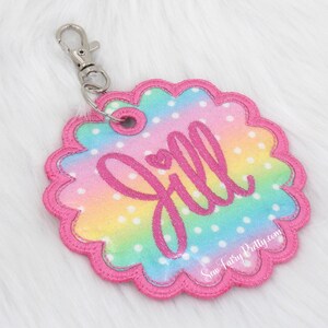 May include: A pink and rainbow-colored fabric keychain with white polka dots and the name "Jill" embroidered in pink. The keychain has a scalloped edge and a silver metal clasp.