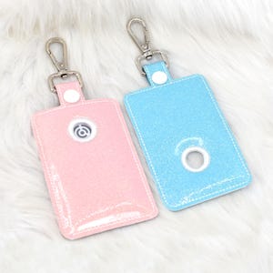 May include: Two glittery rectangular holders with silver-tone hardware. One is light pink, the other is light blue. Each has a silver clasp and a white button. The pink holder has a circular design. The blue holder has a circular opening.