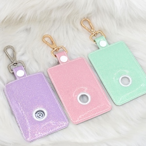 May include: Three glittery dog waste bag holders in lavender, pink, and mint green. Each rectangular holder has a circular opening and a metal clasp for attaching to a leash. The holders are on a white fluffy surface.