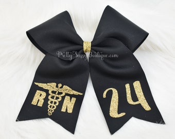 May include: Black hair bow with gold glitter accents. The bow has a gold glitter medical symbol and the letters "RN" on one side and the number "24" on the other side.