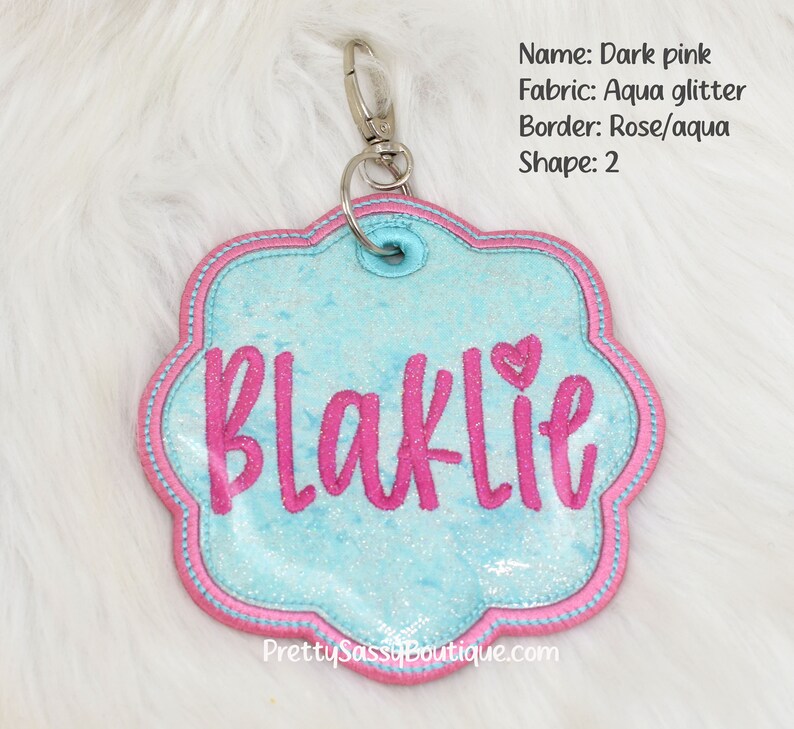 May include: Pink and aqua glitter personalized name tag with the name "Blaklie" in pink lettering. The tag is shaped like a flower and has a pink border.