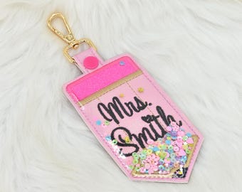 May include: Pink pencil-shaped keychain with glitter and sequins. The keychain has the text "Mrs. Smith" embroidered on it.
