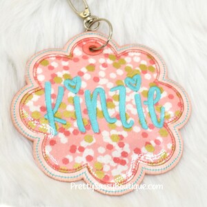May include: Pink and gold glitter flower-shaped keychain with the name "Kinzie" embroidered in turquoise.