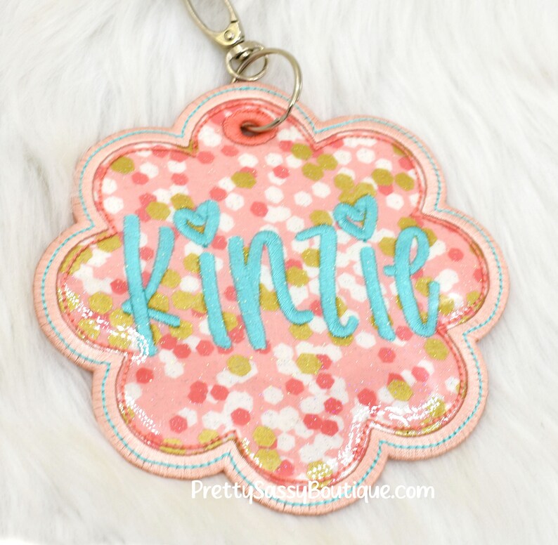 May include: Pink and gold glitter flower-shaped keychain with the name "Kinzie" embroidered in turquoise.