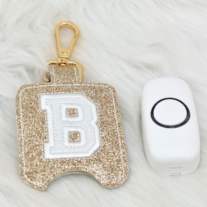 May include: A gold glitter key fob with a white letter "B" and a gold clasp next to a white doorbell with a black circle.