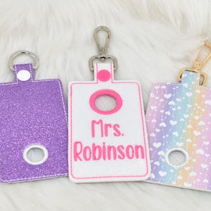 May include: Three different hand sanitizer holders with keychains. The first is purple glitter with a white circle in the center. The second is white with pink text that says "Mrs. Robinson". The third is a rainbow pattern with white hearts.