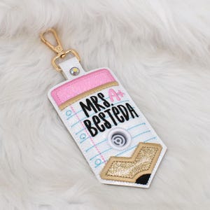May include: A white pencil-shaped keychain with a gold-tone metal clasp. The design features a pink glitter eraser, blue lined paper pattern, and a gold glitter pencil tip. The text "MRS. BESTEDA" is embroidered in black with a pink "A+".