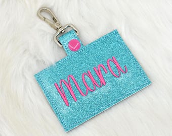 May include: A teal glitter keychain tag with the name "Mara" embroidered in pink script. The tag has a silver clasp and a pink snap closure. The background is a soft, white, fluffy surface.