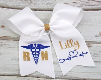May include: White cheer bow with gold glitter accents. One side features a blue medical symbol with the letters "RN" in gold glitter. The other side has the name "Lilly" in gold glitter and a stethoscope with a heart beat graphic.