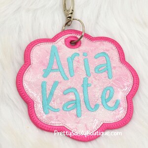 K&ouml;nnte beinhalten: Personalisiertes Namensschild in Pink und T&uuml;rkis mit den Namen "Aria Kate" auf einem pinkfarbenen, glitzernden Hintergrund. Das Schild ist an einem silbernen Schl&uuml;sselring befestigt.