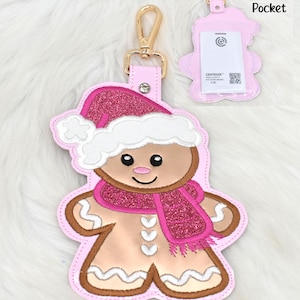 May include: Pink gingerbread man keychain with a gold clasp. The gingerbread man has a pink glittery Santa hat and scarf. The keychain has a clear pocket with the text "Clear Pocket".