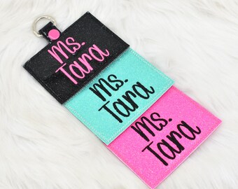 May include: A personalized keychain with three glittery rectangular tags in black, teal, and pink. Each tag features the name "Ms. Tara" in a cursive font. The keychain has a silver ring and a pink accent.