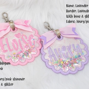 K&ouml;nnte beinhalten: Zwei personalisierte Schl&uuml;sselanh&auml;nger mit Glitzer und Schleifen. Der Schl&uuml;sselanh&auml;nger links ist rosa mit dem Namen "Eloise" und der Schl&uuml;sselanh&auml;nger rechts ist lila mit dem Namen "Avery".