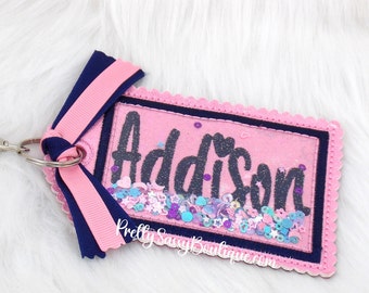 May include: A pink and navy blue keychain tag with the name "Addison" in dark blue script. The tag has a scalloped edge and a clear window filled with sequins. A pink and navy blue ribbon is attached.