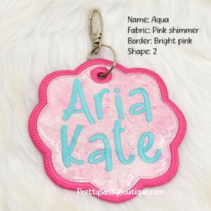 K&ouml;nnte beinhalten: Ein personalisiertes Namensschild aus pinkfarbenem Schimmerstoff mit einem leuchtend pinkfarbenen Rand und einem silbernen Schl&uuml;sselring. Das Schild ist blumenf&ouml;rmig und tr&auml;gt die Namen "Aria Kate", die in t&uuml;rkisfarbenem Garn gestickt sind.