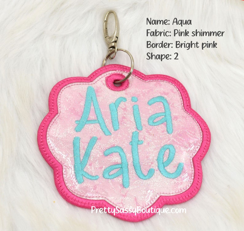 K&ouml;nnte beinhalten: Ein personalisiertes Namensschild aus pinkfarbenem Schimmerstoff mit einem leuchtend pinkfarbenen Rand und einem silbernen Schl&uuml;sselring. Das Schild ist blumenf&ouml;rmig und tr&auml;gt die Namen "Aria Kate", die in t&uuml;rkisfarbenem Garn gestickt sind.