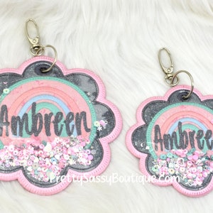 May include: Two pink flower-shaped keychains with a rainbow and glitter inside. The name "Ambreen" is written in black on each keychain.