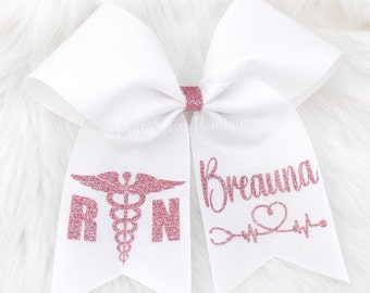 May include: White cheer bow with pink glitter accents. One side features the medical symbol and the letters "RN" and the other side says "Breanna" with a heart and a heartbeat line.