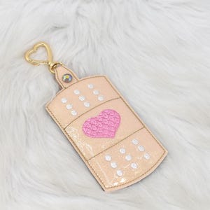 May include: A beige-colored keychain shaped like a bandage, featuring a pink heart and white dot details. It has a gold-colored heart-shaped clasp and a small iridescent gem. The keychain is on a fluffy white surface.
