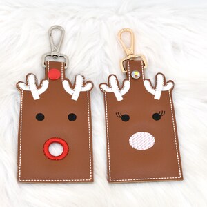 May include: Two brown leather keychains with reindeer faces. Each keychain has white antlers, black eyes, and a red or white nose. The keychains have silver and gold-colored clasps.