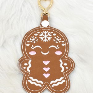 May include: A brown gingerbread man keychain with white stitching and accents. The keychain features a gold-colored heart-shaped clasp. The gingerbread man has pink cheeks, three pink hearts, and snowflake details.
