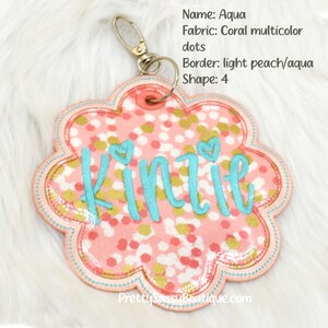 May include: A pink and gold fabric keychain with the name "Kinzie" embroidered in turquoise. The keychain is shaped like a flower with a light peach and aqua border.