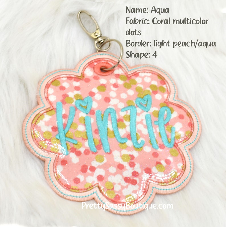 May include: A pink and gold fabric keychain with the name "Kinzie" embroidered in turquoise. The keychain is shaped like a flower with a light peach and aqua border.