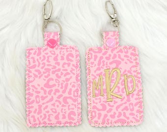 May include: Two pink luggage tags with a leopard print design. One tag has the monogram "MRD" in gold lettering. Both tags have a silver key ring.