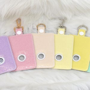 May include: Six pastel-colored pet waste bag holders with metal clip attachments. The holders are rectangular with a circular opening and a white button detail. Colors include lavender, pink, beige, yellow, and mint green.