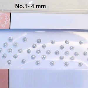Puede incluir: Una caja blanca con un diseño rosa y floral. La caja contiene una hoja de pequeñas gemas redondas, blancas y adhesivas. El texto "No. 1 - 4 mm" está impreso en la caja.