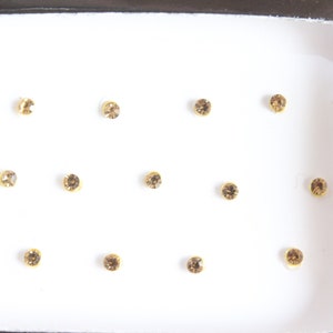 Peut inclure: Un ensemble de douze petits strass ronds dorés dans un récipient en plastique blanc. Les strass sont disposés en grille.